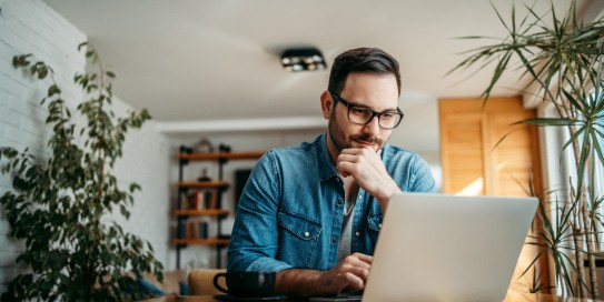 Mann sitzt mit Laptop am Esstisch und ließt interessiert