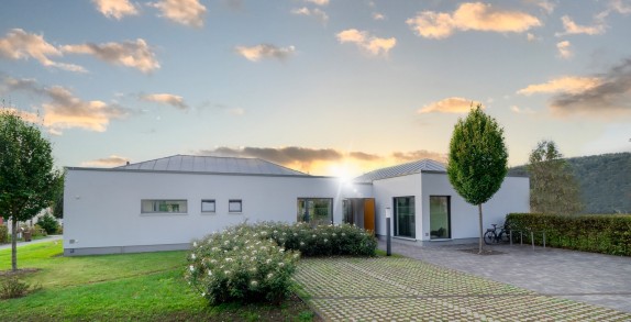 Modernes, ebenerdiges Gebäude mit gepflegtem Vorgarten, gepflasterter Einfahrt und Fahrradständern, im Hintergrund Sonnenuntergang und bewaldete Hügel.