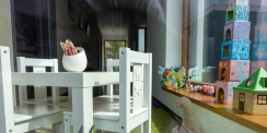Blick durch ein Fenster in eine liebevoll eingerichtete Kinderspielecke mit kleinem weißen Tisch, Stühlen, Spielzeugfiguren, Bauklötzen und Buntstiften.