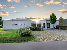 Modernes, ebenerdiges Gebäude mit gepflegtem Vorgarten, gepflasterter Einfahrt und Fahrradständern, im Hintergrund Sonnenuntergang und bewaldete Hügel.