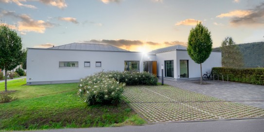 Modernes, ebenerdiges Gebäude mit gepflegtem Vorgarten, gepflasterter Einfahrt und Fahrradständern, im Hintergrund Sonnenuntergang und bewaldete Hügel.