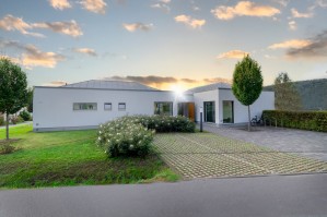 Modernes, ebenerdiges Gebäude mit gepflegtem Vorgarten, gepflasterter Einfahrt und Fahrradständern, im Hintergrund Sonnenuntergang und bewaldete Hügel.