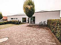 Moderner Eingangsbereich eines Gebäudes mit gepflastertem Vorplatz, Baum in der Mitte, Fahrradständer und gepflegter Hecke.