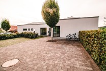 Moderner Eingangsbereich eines Gebäudes mit gepflastertem Vorplatz, Baum in der Mitte, Fahrradständer und gepflegter Hecke.