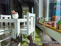 Blick durch ein Fenster in eine liebevoll eingerichtete Kinderspielecke mit kleinem weißen Tisch, Stühlen, Spielzeugfiguren, Bauklötzen und Buntstiften.