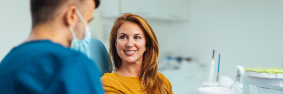 Zahnärztlicher Mitarbeiter im Gespräch mit einer Patientin auf dem Behandlungsstuhl, die lächelnd zuhört.