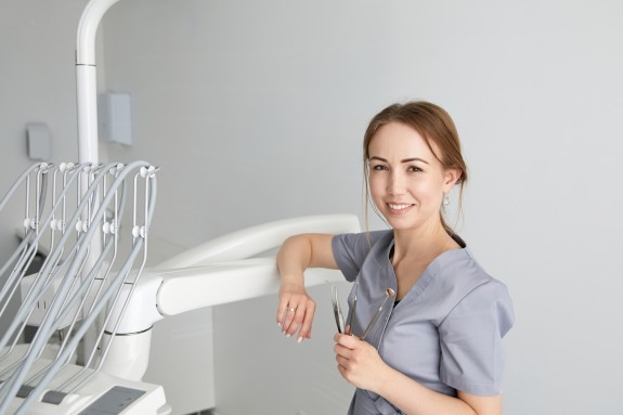 Zahnmedizinische Fachangestellte in grauer Kleidung steht in einem Behandlungsraum und hält zahnärztliche Instrumente in der Hand.